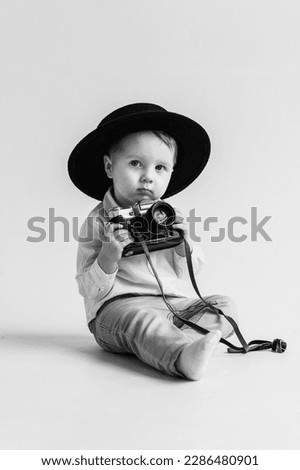 Child in hat sitting on floor and play with photo camera. World Photographers Day. Fun kid holds retro vintage camera isolated on white wall. Children's studio portrait. Mockup. Black and white photo.