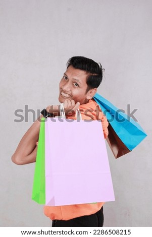 portrait Asian adult man wearing orange t-shirt carrying grocery paper bag and looking at camera in funny pose. pink background screen