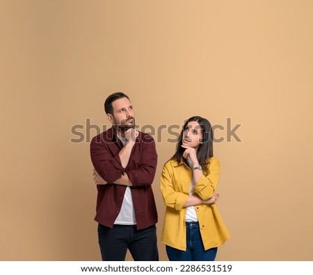 Uncertain young couple with hands on chins thinking for solution. Serious young man and woman dressed in shirts contemplating and looking up while standing over beige background Royalty-Free Stock Photo #2286531519