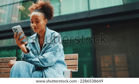 Cute girl with ponytail, wearing denim jacket, in crop top with national pattern, sitting on bench and using smartphone on modern buildings background. Royalty-Free Stock Photo #2286538649