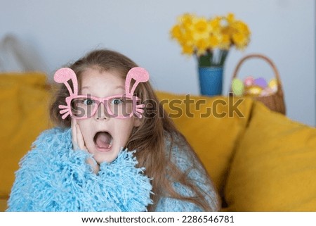 Cute little child girl  in blue fluffy coat wearing bunny ears glasses a on Easter day. Easter girl portrait, funny emotions, surprise.	
