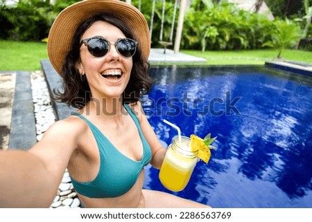 Happy woman in a bikini drinking juice taking selfie in the swimming pool during summer vacation in tropical resort.