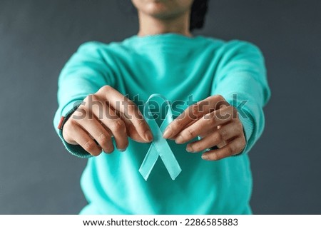 Close-up teal awareness ribbon holded by woman hands to support cancer survivor. Ovarian Cancer month, cervical cancer day.  Royalty-Free Stock Photo #2286585883