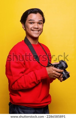 Side view of young photographer standing confidently and smiling while holding camera and  isolated on yellow color background