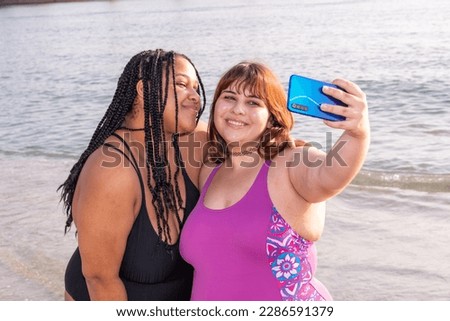 Two cute plus size girls in bikinis take photos on the beach. Young women take selfies with their smartphone. Couple of women use their phone to take vacation photos
