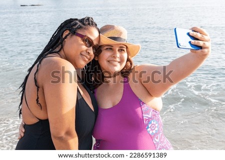 Two cute plus size girls in bikinis take photos on the beach. Young women take selfies with their smartphone. Couple of women use their phone to take vacation photos