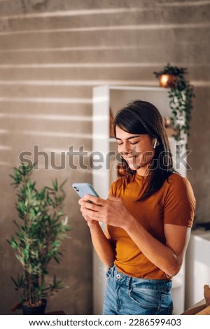 Beautiful woman checking social media while using smartphone and airpods at home. Smiling young female using mobile phone app and playing game, shopping online or reading news. Copy space. Royalty-Free Stock Photo #2286599403