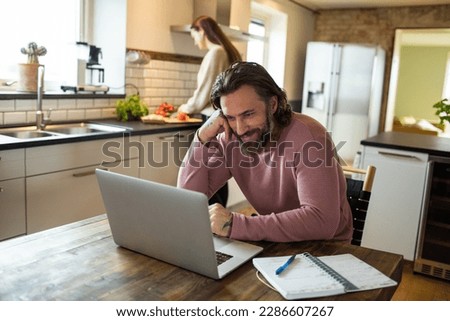 Young adult man using a laptop while his wife is working in the kitchen behind him