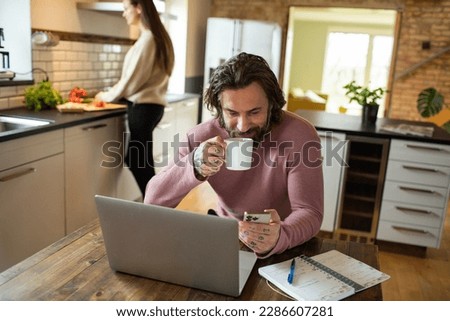 Young adult man using a smart phone while his wife is working in the kitchen behind him