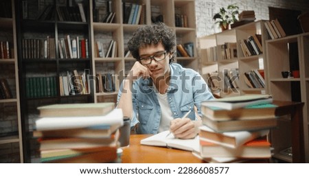Asian student with curly hair is in library, sits at desk full of books. Guy wearing glasses preparing for exam and studying - education concept close up 