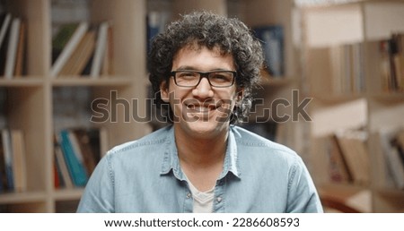 Asian student with curly hair is standing in library. Guy wearing glasses looks at camera and happily smiles - portrait close up 