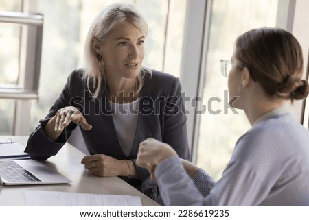 Two young and mature business colleagues women talking at work table, discussing project strategy, plan. Mature professional speaking to coworker, training intern, new employee Royalty-Free Stock Photo #2286619235