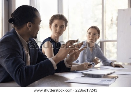 African millennial businessman talking to colleagues on office meeting, presenting idea for project, speaking, telling marketing strategy, explaining plan, task, sitting at work table with coworkers Royalty-Free Stock Photo #2286619243