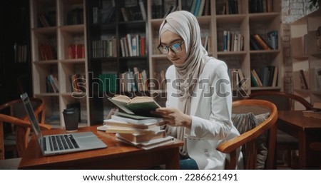 Female muslim student is studying at desk in library, using laptop and books. Girl wearing hijab is preparing for exams - student lifestyle, modern Islam concept close up
