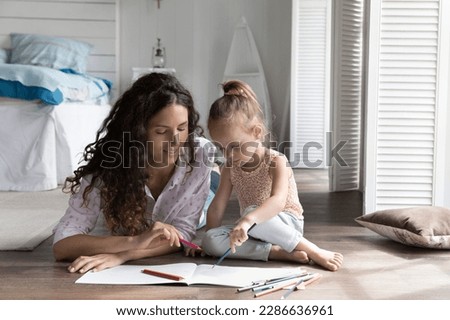 Mom engaging little kid into creative hobby, resting on heating floor at home, drawing in colorful pencils in paper album, talking, spending family leisure, weekend with daughter girl