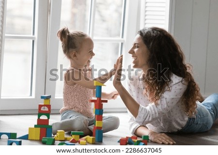 Cheerful mom and kid girl clapping hands, giving high five over colorful toy towers stacked from wooden constructing blocks, playing on heating floor, celebrating successful building Royalty-Free Stock Photo #2286637045