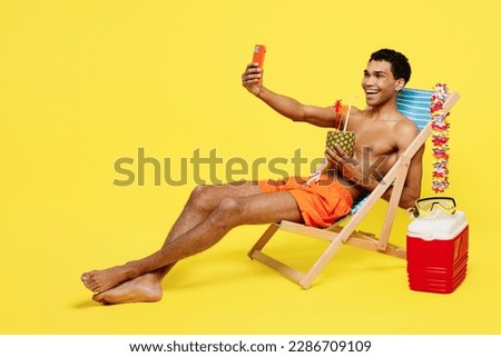 Full body young man wear shorts swimsuit relax near hotel pool sit in deckchair do selfie shot on mobile cell phone drink cocktail isolated on plain yellow background. Summer sea rest sun tan concept