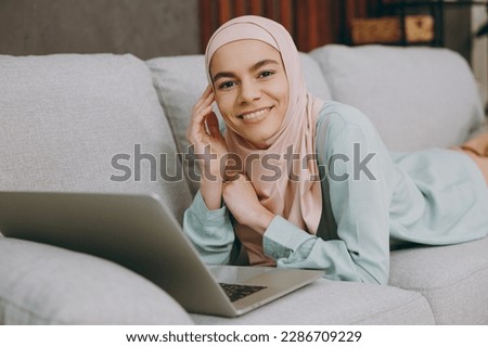 Young happy muslim IT woman wear hijab casual clothes use work on laptop pc computer sit on sofa couch stay at home flat rest relax spend free spare time in living room indoor People uae islam concept