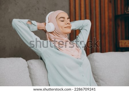 Young fun muslim woman wears hijab casual clothes headphones listen music sit on sofa couch stay at home flat rest relax spend free time in living room indoor People middle eastern uae islam concept