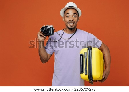 Traveler black man wear purple t-shirt hat hold suitcase photo camera isolated on plain orange color background. Tourist travel abroad on weekends spare time getaway. Air flight trip journey concept