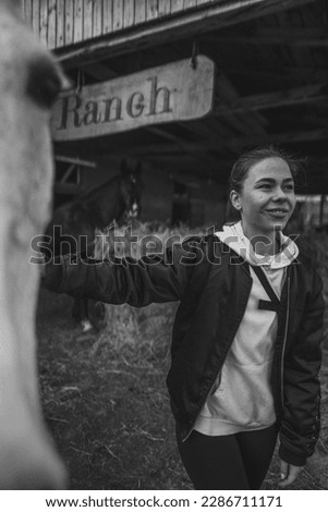 A small woman and a large tall horse on the ranch. The black and white horse is very beautiful and friendly. Blurred wooden background and warm colours of the picture. The sky is cloudy but sun shines