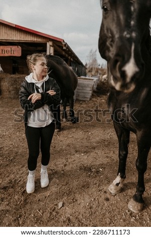 A small woman and a large tall horse on the ranch. The black and white horse is very beautiful and friendly. Blurred wooden background and warm colours of the picture. The sky is cloudy but sun shines