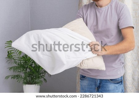 A man tucks a pillow with a fresh bright white pillowcase. Making the bed with fresh bed linen by a white man. The day of the change of bed linen. The day of washing bed linen in the laundry room. Royalty-Free Stock Photo #2286721343