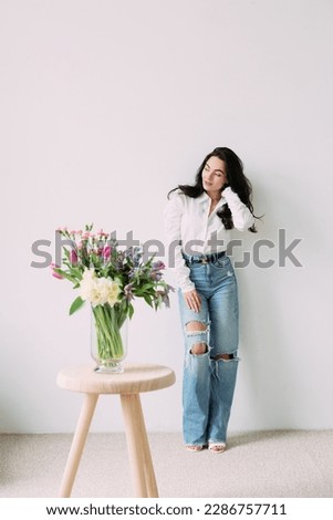 Young pensive woman standing near wall next to beautiful bouquet of flowers on the table. Mothers day, birthday, anniversary, celebration, holiday, greeting, festive event.