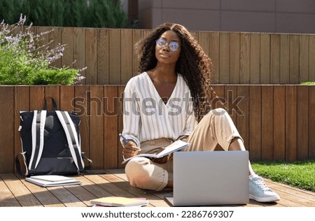 Young pretty black African woman university student learning online using laptop computer, taking notes, studying outdoors sitting outside uni campus area looking at camera.