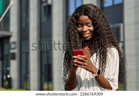 Happy young African woman model using smartphone standing outdoors. Beautiful black woman holding cell phone doing online shopping, checking apps looking at cellphone technology device in city.