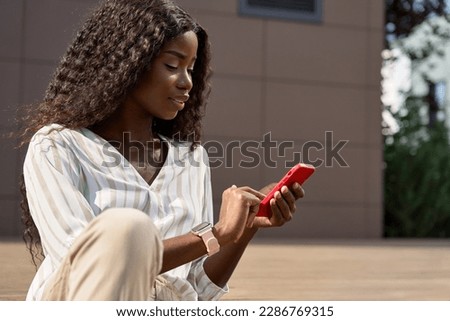 Pretty young African woman model using smartphone sitting outdoors. Beautiful black woman holding cell phone doing online shopping, looking at cellphone technology device sitting outside in city.
