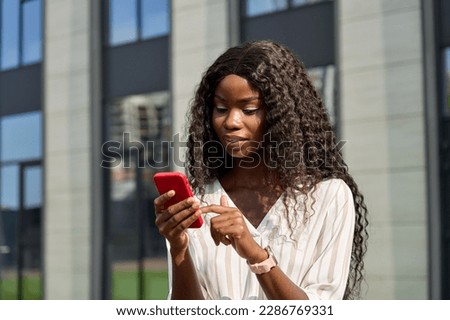 Young African model using smartphone standing outdoors. Pretty black business woman holding cell phone doing online shopping, scrolling, browsing looking at cellphone technology device in city.
