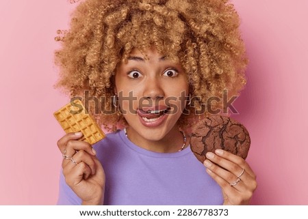 Headshot of curly haired young European woman holds tasty waffle and chocolate cookie licks lips wants to eat sweet appetizing food dressed in casual purple t shirt isolated over pink background Royalty-Free Stock Photo #2286778373