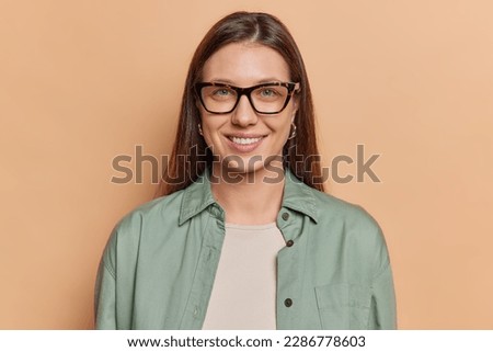 Portrait of lovely young European woman with dark straight hair smiles pleasantly looks through transparent eyeglasses dressed in shirt expresses positive emotions isolated over brown background