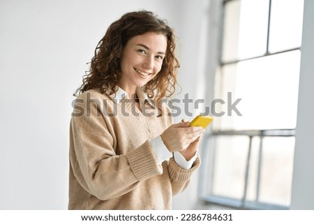 Happy young woman holding smartphone standing indoors at home using mobile cell phone, buying online in ecommerce shop, texting messages on cellphone, browsing websites or downloading new apps.