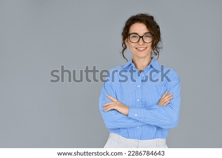 Happy young smiling confident professional business woman wearing blue shirt and glasses, happy pretty female executive looking at camera, standing arms crossed isolated at gray background, portrait.
