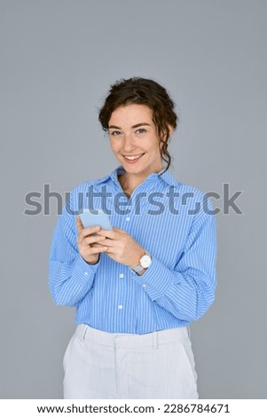 Happy young business woman manager holding smartphone standing at gray wall using mobile cell phone, checking apps, browsing online on cellphone typing on cellular technology device. Royalty-Free Stock Photo #2286784671