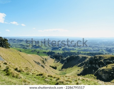 The picture shows a panoramic view from the top of Te Mata Peak over green hills and meadows of Hawke's Bay, New Zealand on a sunny day with a blue sky. 