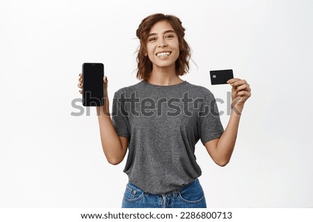 Portrait of girl posing with mobile phone and credit card, showing smartphone app screen, interface, standing over white background.