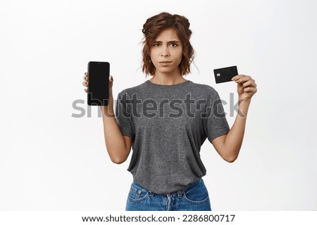 Confused young woman shows black credit card and smartphone screen, girl with doubts looking at camera while showing mobile phone app, white background.