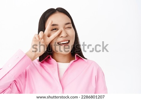 Close up of asian girl shows peace sign near face, winks at you and smiles, stands in pink shirt over white background. Copy space