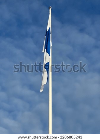 There is a Finnish flag on the flag pole. In the picture, the flag is against the blue sky. There are misty clouds in the sky. The air is calm and the flag is not blowing with the wind.