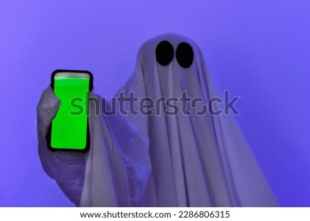 The ghost shows a phone with a green screen. Phone with chromakey. The ghost makes an order from smartphone on Halloween. Free space. The ghost poses with the phone.