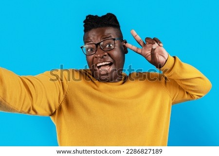Cheerful Black Guy In Eyeglasses Showing Peace Gesture At Camera While Taking Selfie, Happy Excited Young African American Man Posing At Camera Over Blue Background, Demonstrating V-Sign