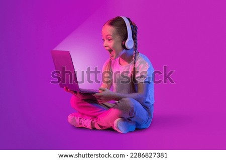 Amazed shocked cute preteen blonde girl with two braids using laptop with shining screen and wireless headphones, playing video game or watching content online, colorful background with neon light