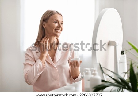 Beautiful middle aged woman applying moisturizing cream on her neck while sitting near mirror at home, attractive mature female enjoying domestic beauty routine, holding jar with nourishing lotion