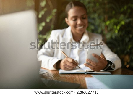Happy millennial african american businesswoman in white suit reading message on smartphone, uses laptop in eco cafe with green plants interior. App for business, work with gadget remotely
