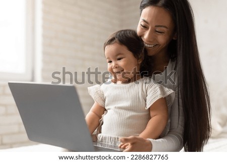 Happy Asian Mom And Little Baby Daughter Using Laptop Computer Watching Cartoons Online And Browsing Internet Sitting In Bedroom At Home. Modern Children And Gadgets Concept