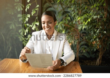 Cheerful millennial african american businesswoman, ceo in white suit reading document, work in cafe office with green plants interior. Modern business, startup, advertising and offer