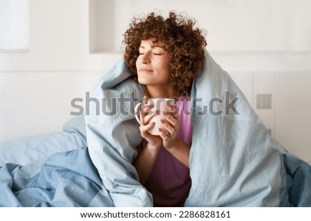 Relaxed curly woman with closed eyes wrapped in blanket while holding mug of hot beverage on bed in weekend Royalty-Free Stock Photo #2286828161